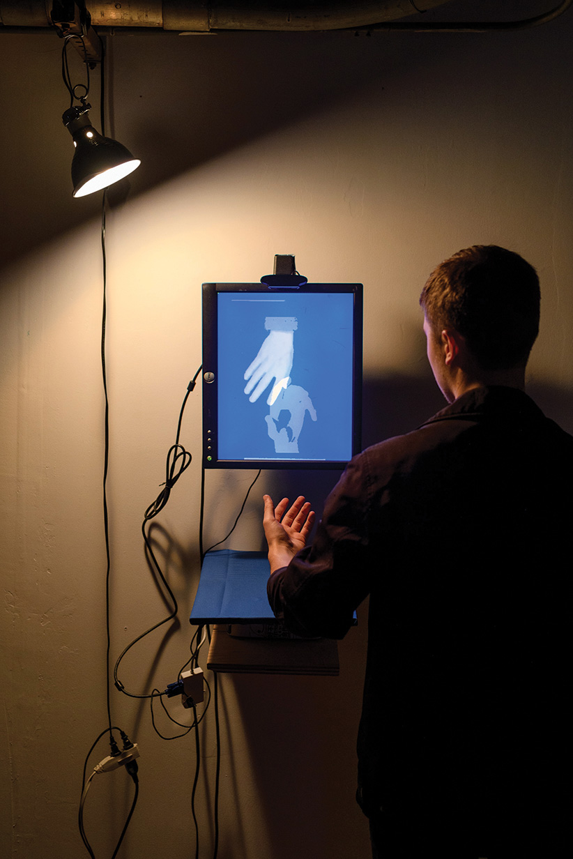 A wide shot of the installation, showing it's messy wiring underneath a warm lamp. The lamp casts light on a viewer who is interacting with the installation.